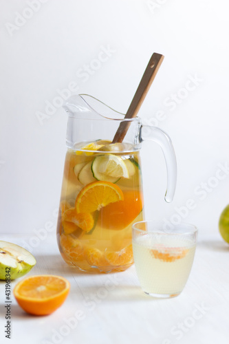 Fruit lemonade with fresh orange, cucumber, tangerine, lemon, apple and honey in glass jug with wooden spoon on white background