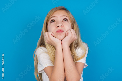 Portrait of sad beautiful Caucasian little girl wearing white T-shirt over blue background hands face look empty space