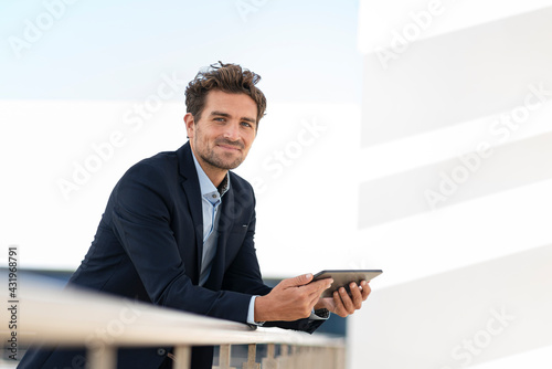 Smiling businessman holding digital tablet while standing by railing at office terrace photo