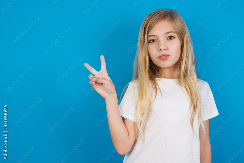 beautiful Caucasian little girl wearing white T-shirt over blue background makes peace gesture keeps lips folded shows v sign. Body language concept beautiful Caucasian little girl wearing white T-shirt over blue background makes peace gesture keeps lips folded shows v sign. Body language concept