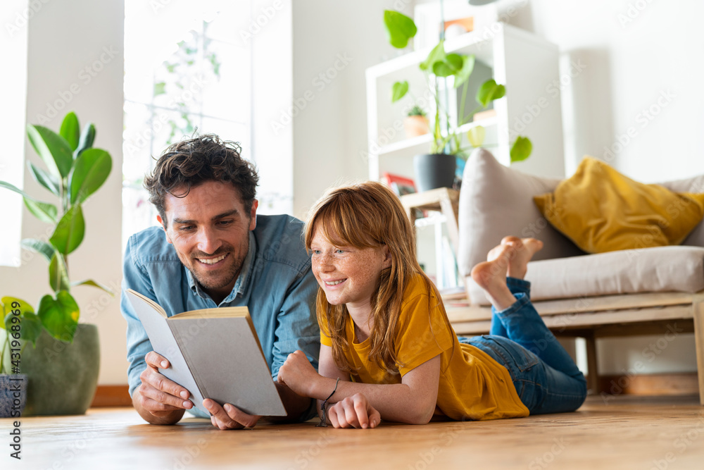 Smiling father and daughter reading book together while lying down on floor at home Smiling father and daughter reading book together while lying down on floor at home