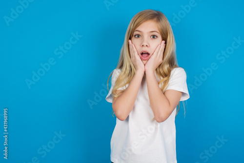 Upset beautiful Caucasian little girl wearing white T-shirt over blue background touching face with two hands
