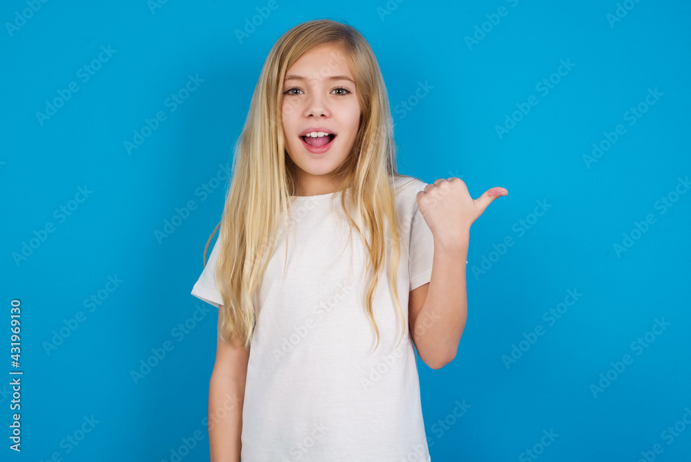 Impressed beautiful Caucasian little girl wearing white T-shirt over blue background point back empty space Impressed beautiful Caucasian little girl wearing white T-shirt over blue background point back empty space
