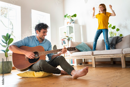 Father playing guitar while daughter dancing on sofa in living room at home