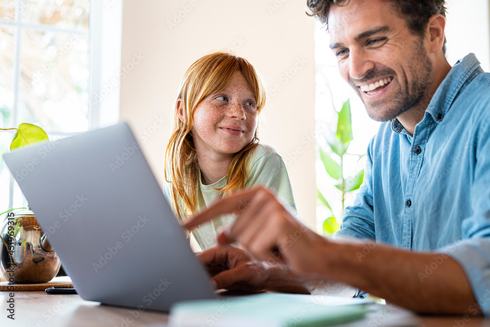 Smiling redhead girl looking at father working on laptop in living room Smiling redhead girl looking at father working on laptop in living room
