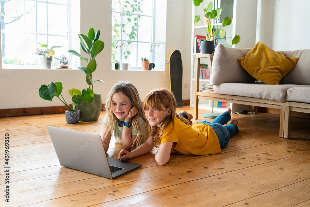 © steve brookland/Westend61 - Smiling female friends looking at laptop while lying on front in living room