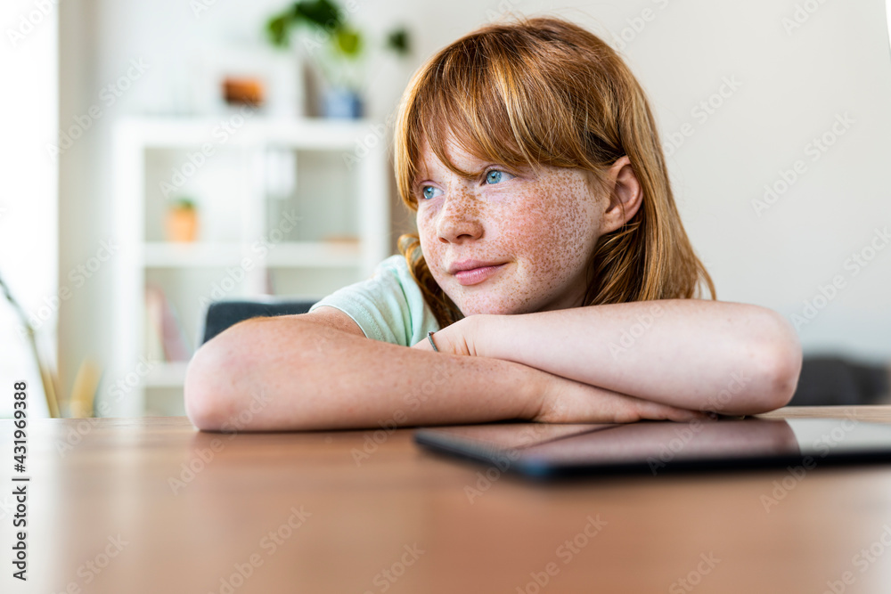 Thoughtful redhead girl looking away while sitting at table in living room Thoughtful redhead girl looking away while sitting at table in living room