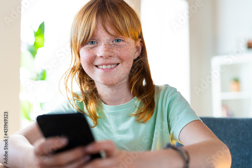 Cute smiling redhead girl holding smart phone at home