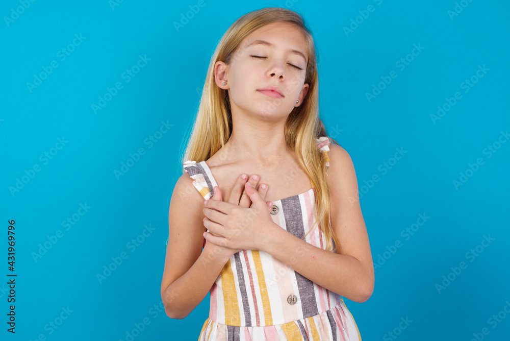 beautiful Caucasian little girl wearing stripped dress against blue wall closes eyes and keeps hands on chest near heart, expresses sincere emotions, being kind hearted and honest. beautiful Caucasian little girl wearing stripped dress against blue wall closes eyes and keeps hands on chest near heart, expresses sincere emotions, being kind hearted and honest.