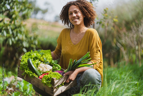 Smiling woman squatting while holding crate of fresh vegetables in garden