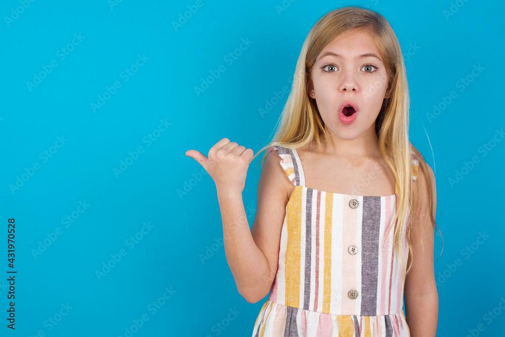 Shocked beautiful Caucasian little girl wearing stripped dress over blue background points with thumb away, indicates something. Check this out. Advertisement concept. Shocked beautiful Caucasian little girl wearing stripped dress over blue background points with thumb away, indicates something. Check this out. Advertisement concept.