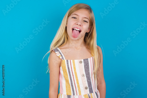 beautiful Caucasian little girl wearing stripped dress over blue background with happy and funny face smiling and showing tongue.