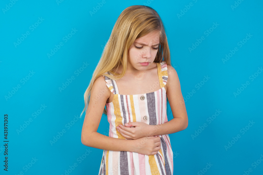beautiful Caucasian little girl wearing stripped dress over blue background suffering from strong stomachache.