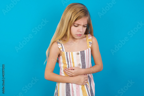 beautiful Caucasian little girl wearing stripped dress over blue background suffering from strong stomachache.