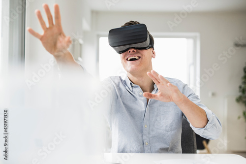Cheerful male professional wearing wearing virtual reality simulator while sitting at desk in home office photo