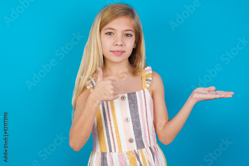 Happy cheerful beautiful Caucasian little girl wearing stripped dress over blue background showing thumb up and pointing with the other hand. I recommend this.