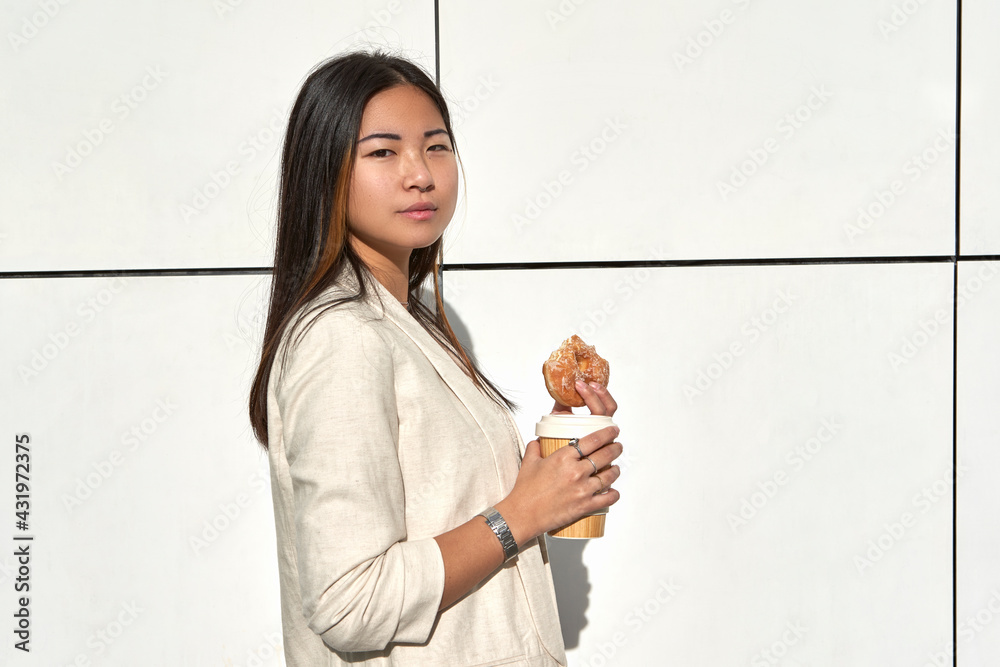 Confident young woman standing with reusable cup and doughnut against wall Confident young woman standing with reusable cup and doughnut against wall
