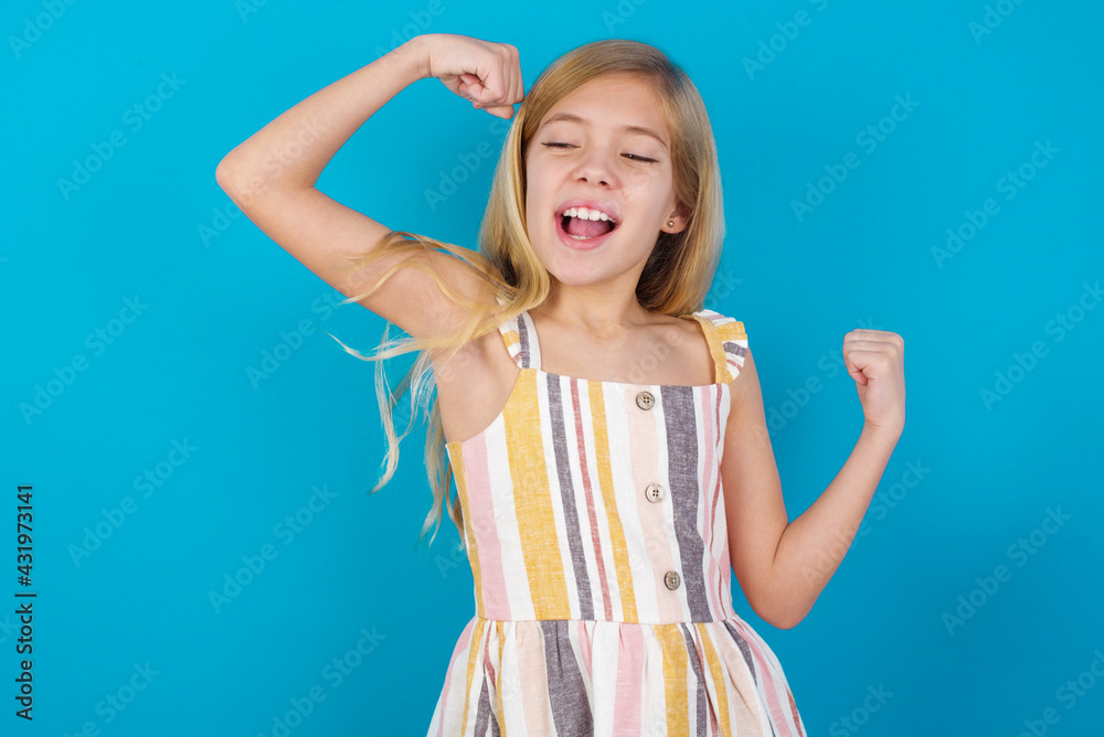 Yes I am winner. Portrait of charming delighted and excited beautiful Caucasian little girl wearing stripped dre raising up fist in triumph and victory smiling achieving success grinning from delight.