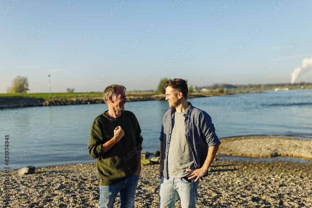 Cheerful father and son discussing while standing against river on sunny day Cheerful father and son discussing while standing against river on sunny day