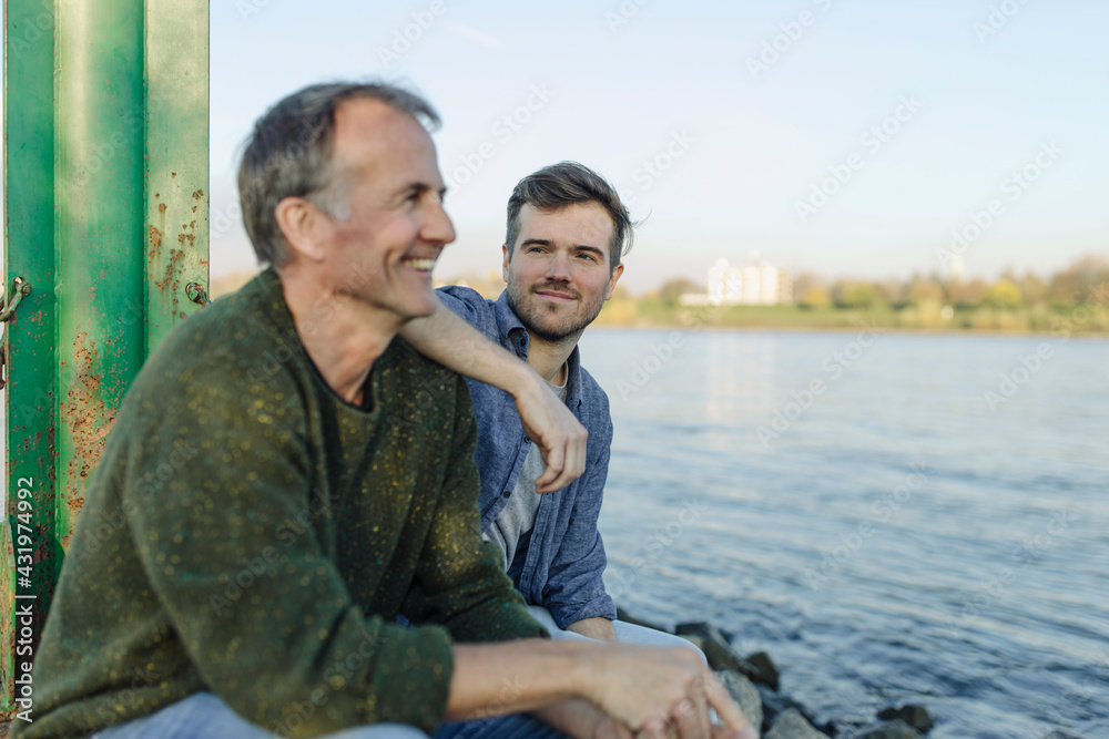 Smiling son looking at happy father against river and sky Smiling son looking at happy father against river and sky
