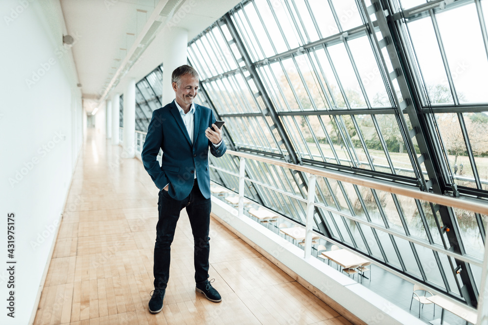 Smiling businessman with hand in pocket using mobile phone while standing in office corridor Smiling businessman with hand in pocket using mobile phone while standing in office corridor