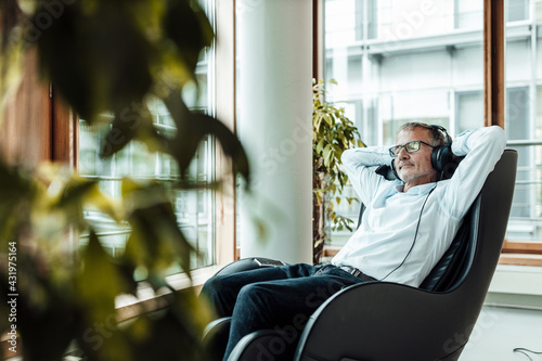 Relaxed businessman with hands behind head listening music through headphones while sitting on massage chair in office corridor photo