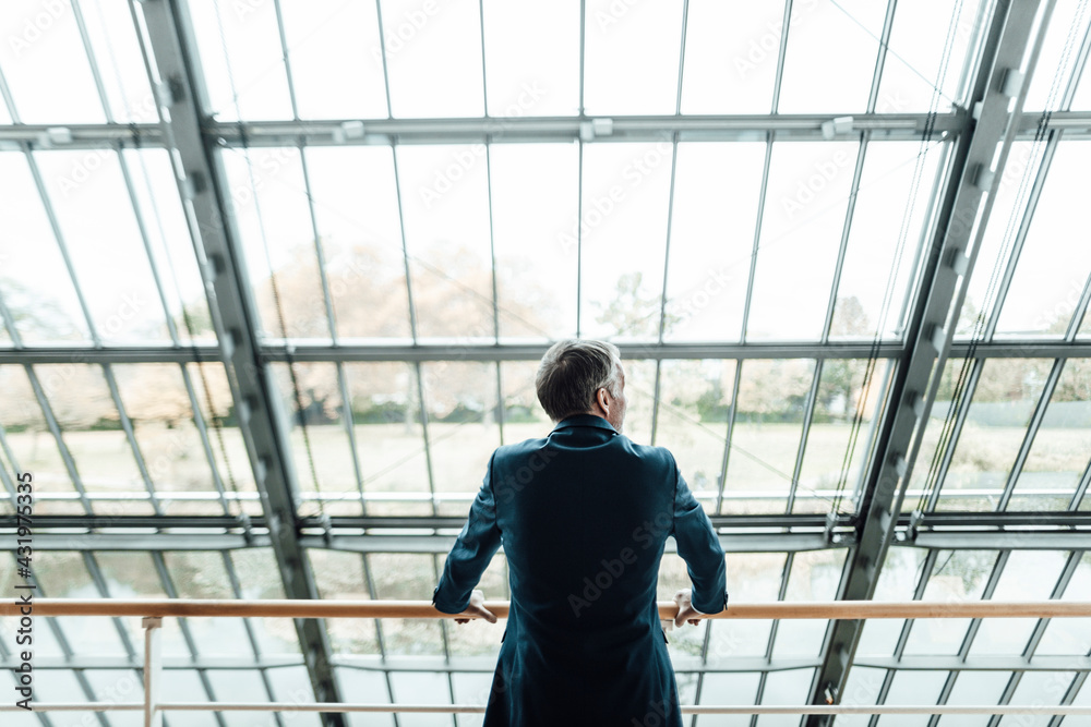Male entrepreneur looking through window while leaning on railing in office corridor Male entrepreneur looking through window while leaning on railing in office corridor
