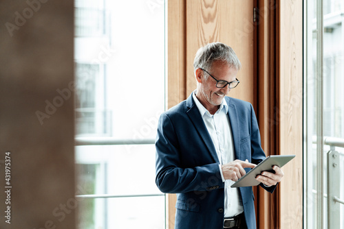 Smiling businessman using digital tablet while standing against glass window