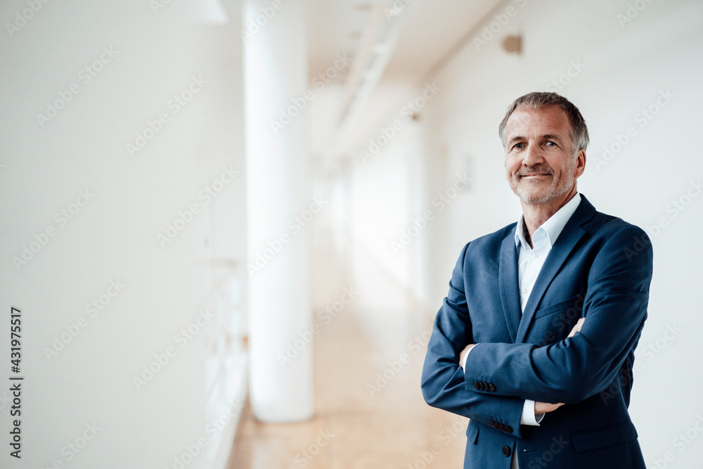 Smiling senior male entrepreneur with arms crossed standing against wall in office corridor Smiling senior male entrepreneur with arms crossed standing against wall in office corridor