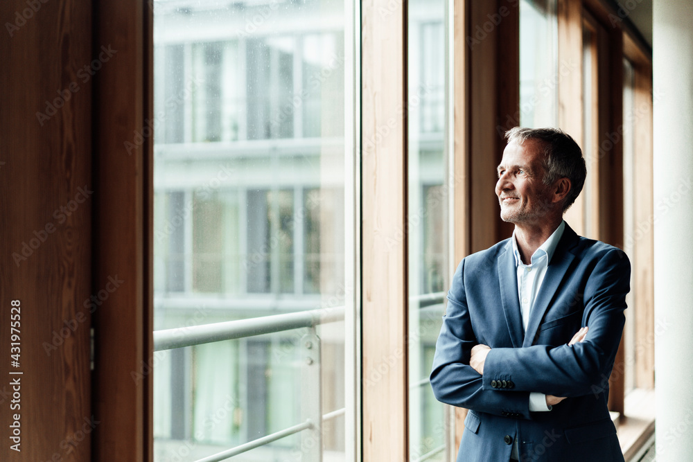 Contemplating businessman looking away while standing with arms crossed at window in office Contemplating businessman looking away while standing with arms crossed at window in office