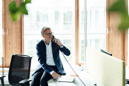 Smiling male business professional with laptop looking away while talking through telephone on desk