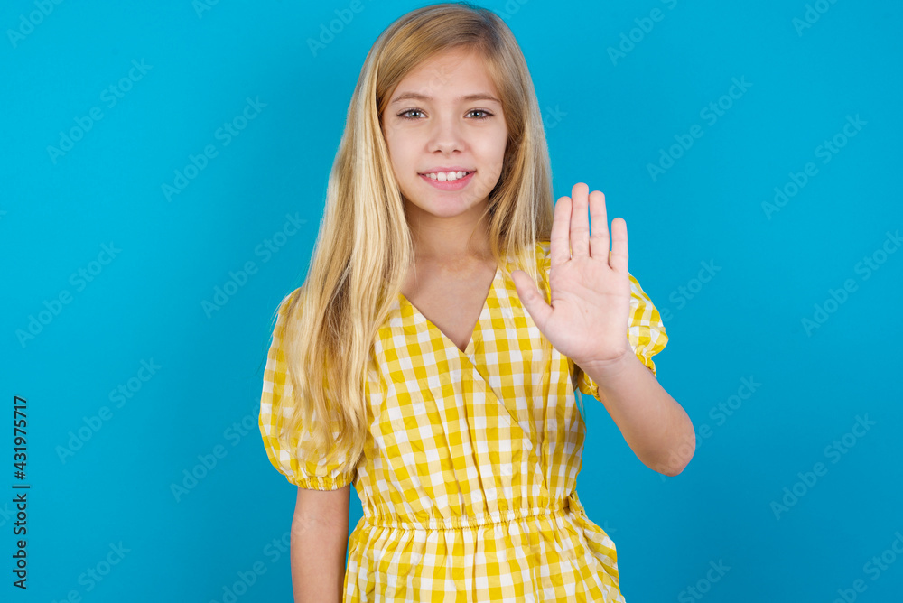 beautiful Caucasian little girl wearing yellow dress over blue background shows stop sign prohibition symbol keeps palm forward to camera with strict expression beautiful Caucasian little girl wearing yellow dress over blue background shows stop sign prohibition symbol keeps palm forward to camera with strict expression