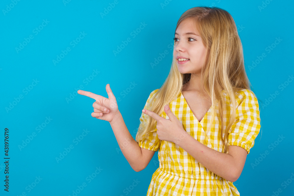 Optimistic beautiful Caucasian little girl wearing yellow dress over blue background points with both hands and looking at empty space. Optimistic beautiful Caucasian little girl wearing yellow dress over blue background points with both hands and looking at empty space.