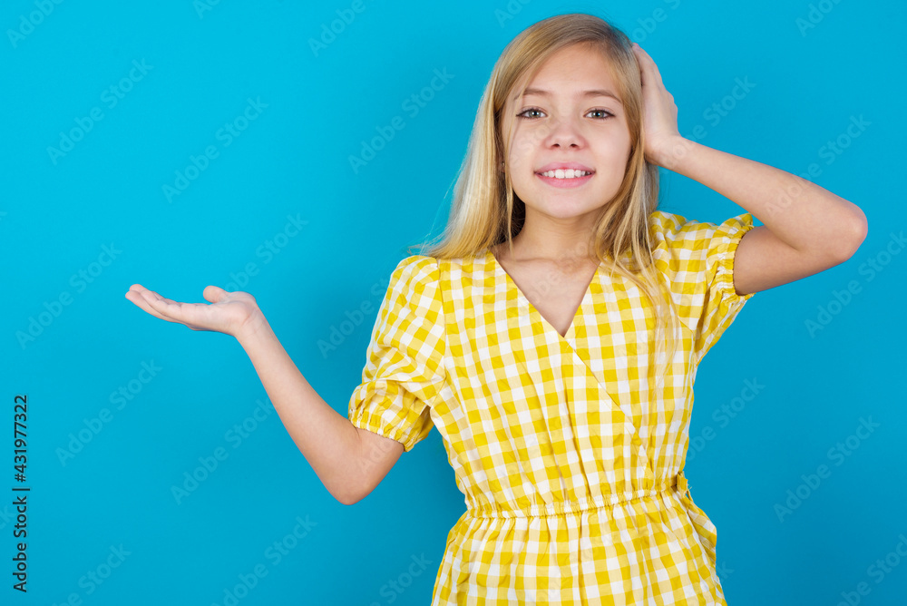 Fototapeta premium Shocked amazed surprised beautiful Caucasian little girl wearing yellow dress over blue background hold hand offering proposition