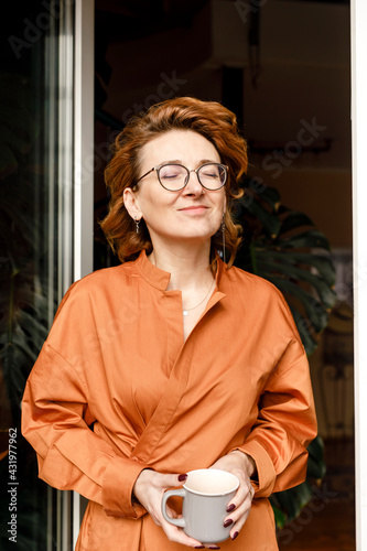 Mature woman with eyes closed holding coffee cup while standing against door