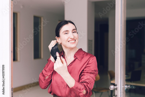 Smiling businesswoman removing protective face mask while standing at glass door during pandemic