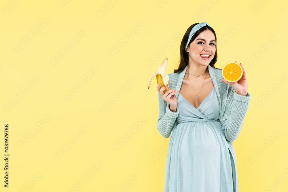 happy pregnant woman smiling at camera while holding banana and juicy orange isolated on yellow. happy pregnant woman smiling at camera while holding banana and juicy orange isolated on yellow.