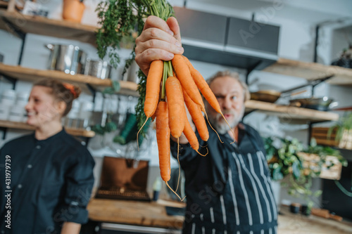 Chef holding organic carrot while standing with colleague at kitchen photo