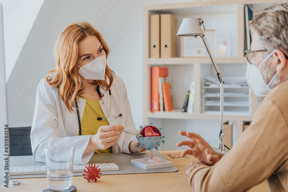 Female doctor with protective face mask explaining over artificial Coronavirus model to patient while sitting at office Female doctor with protective face mask explaining over artificial Coronavirus model to patient while sitting at office