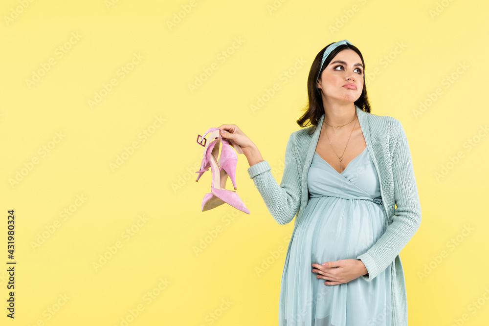 thoughtful pregnant woman holding high heeled shoes isolated on yellow. thoughtful pregnant woman holding high heeled shoes isolated on yellow.