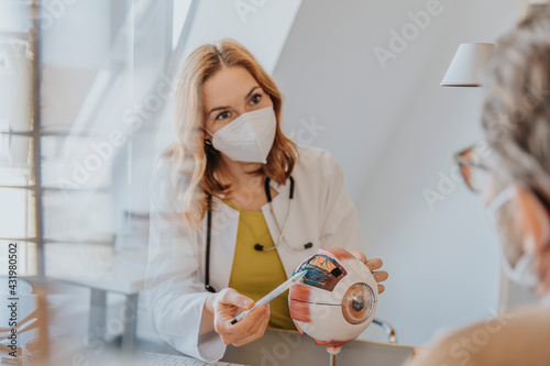 Doctor with protective face mask explaining patient over eye model at clinic
