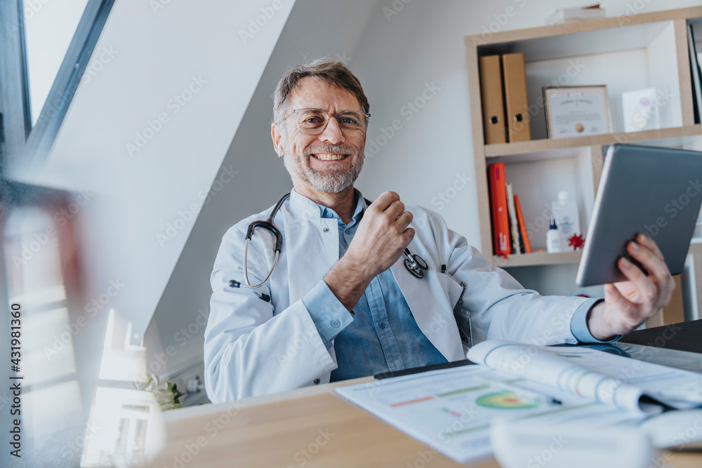 Smiling male doctor with digital tablet sitting by desk at doctor's office Smiling male doctor with digital tablet sitting by desk at doctor's office