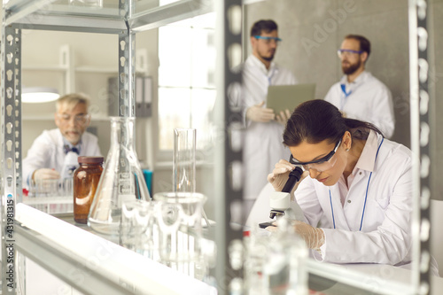 Science research laboratory workflow. Focus on woman scientist wearing safety goggles and lab coat working with microscope equipment analyzing test sample after chemical experiment