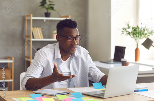 Portrait of young cheerful african american man at home office desk looking at laptop screen having video call on computer. Business training webinar, e-learning, video-conference with team mates