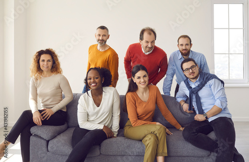 Group portrait of happy confident businessmen and businesswomen on couch in new company office. Diverse team of business partners and colleagues sitting or standing by gray sofa and smiling at camera