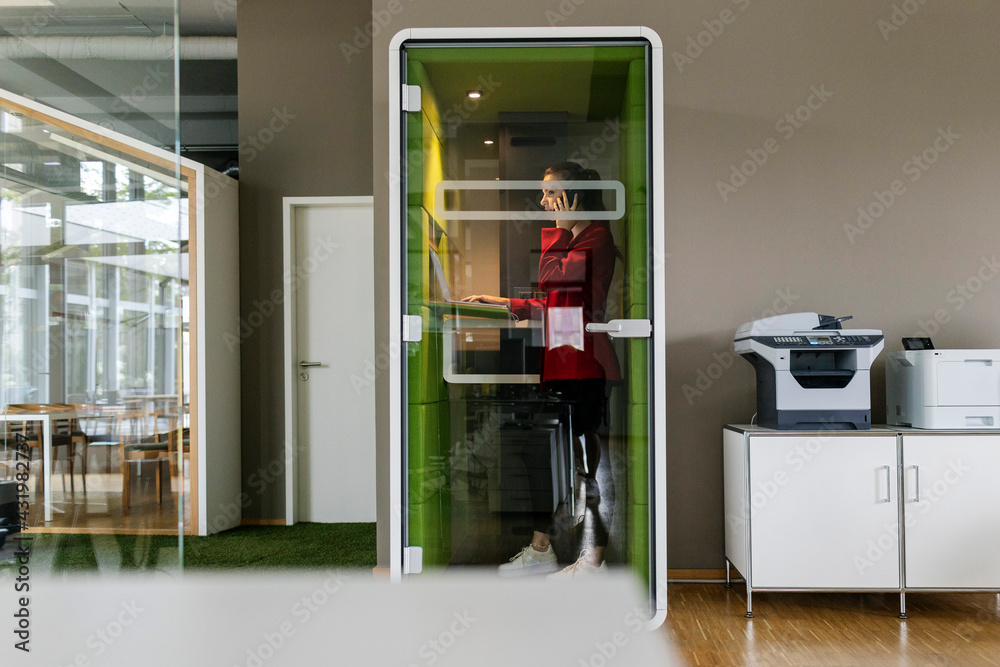 Female entrepreneur talking on smart phone while standing in soundproof cabin at office Female entrepreneur talking on smart phone while standing in soundproof cabin at office