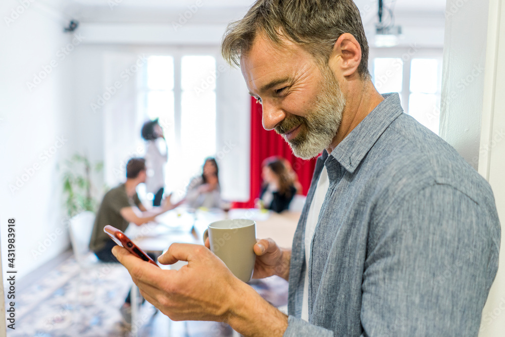 Smiling male entrepreneur using smart phone during coffee break in office Smiling male entrepreneur using smart phone during coffee break in office