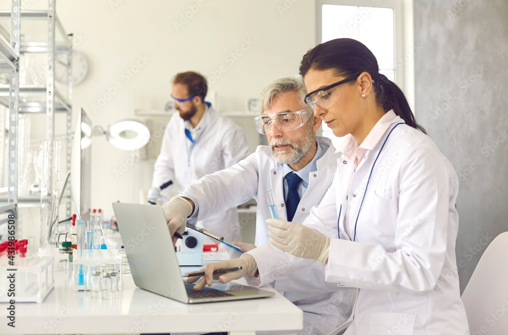 Old professor and young scientist with analysis sample working at laboratory discussing test result looking at on laptop screen. Chemistry research, new drug vaccine discovery concept Old professor and young scientist with analysis sample working at laboratory discussing test result looking at on laptop screen. Chemistry research, new drug vaccine discovery concept