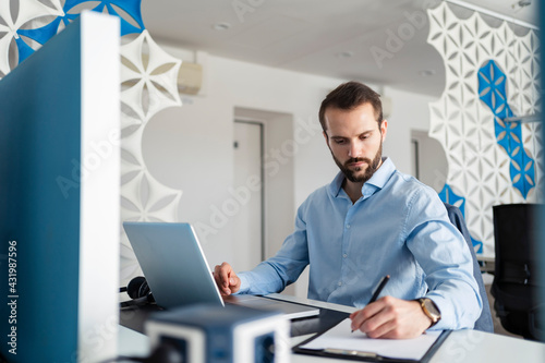 Young professional writing in paper while sitting with laptop at office