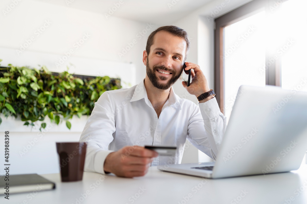 Smiling businessman with credit card and laptop talking on mobile phone at office Smiling businessman with credit card and laptop talking on mobile phone at office